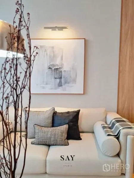 Condominium for rent or sale in Khlong Toei, Bangkok - Close‑up of plush sofa with layered cushions beneath abstract artwork and soft wall lighting.