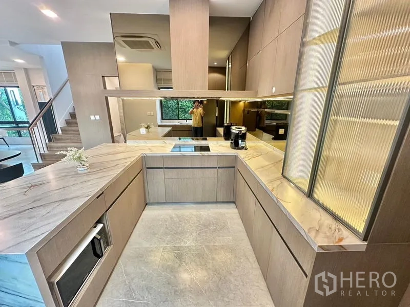 Detached House for rent in Samut Prakan - U-shaped kitchen with marble-look countertops, mirrored backsplash and built-in appliances.