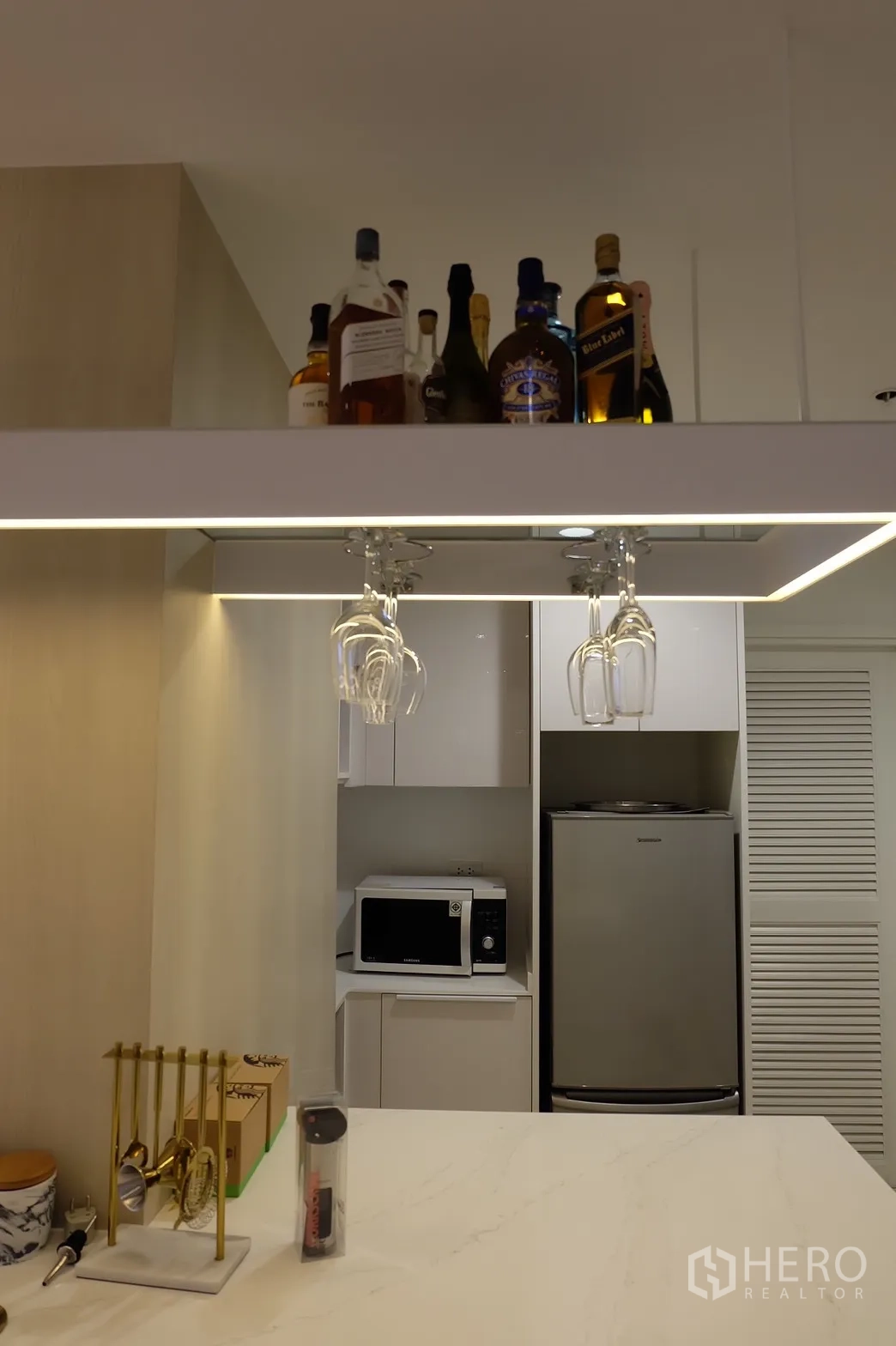 Condominium for rent in Bang Rak, Bangkok - Close-up of illuminated bar shelf with bottles and hanging glasses above the kitchen island.