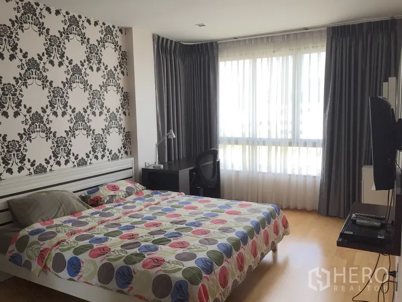 Condominium for rent or sale in Khlong Toei, Bangkok - Primary bedroom with patterned feature wall, large window, work desk and wall-mounted TV.