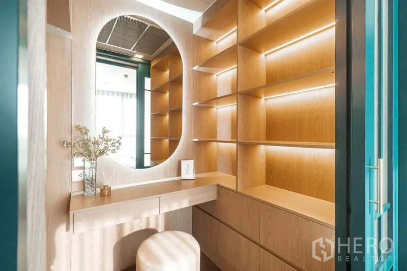 Condominium for rent in Khlong Toei, Bangkok - Bright dressing corner with an oval mirror, stool, and wood shelves illuminated by LED strips.