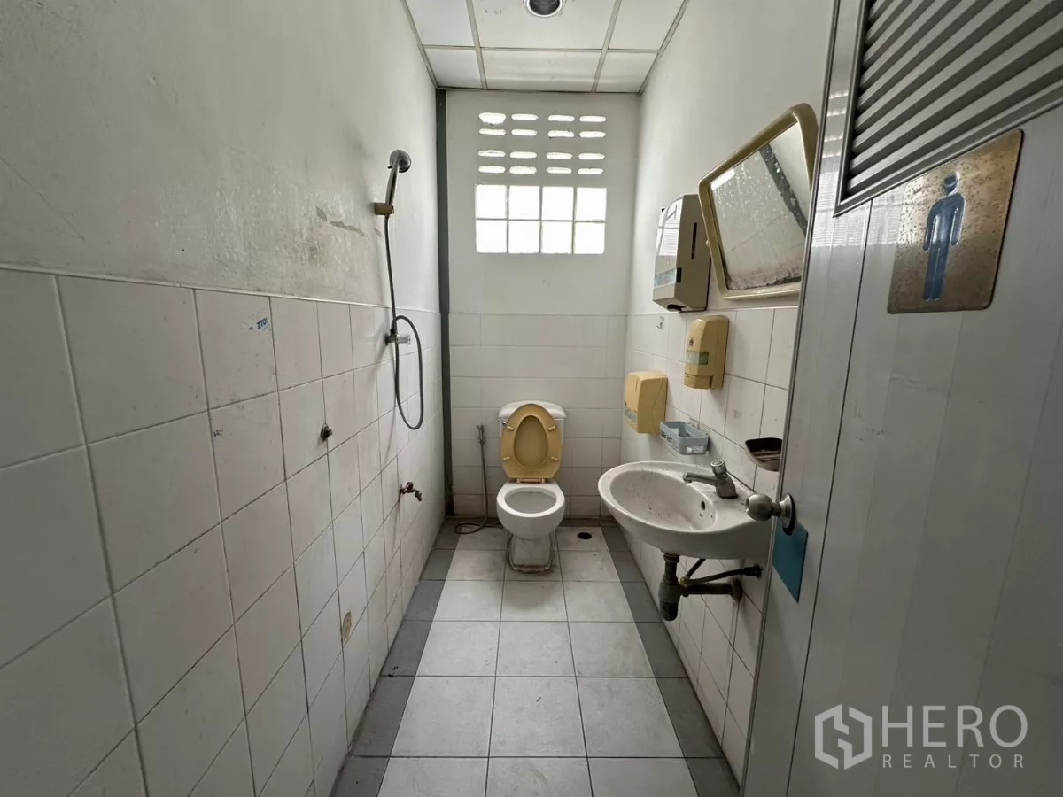 Warehouse for rent in Suan Luang, Bangkok - Men’s restroom with white tiles, toilet, sink, mirror, and handheld shower.