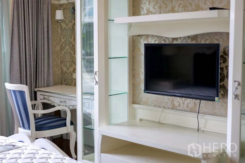 Condominium for rent in Khlong Toei, Bangkok - Bedroom corner with white shelving unit, wall-mounted TV, and study desk against patterned wallpaper.