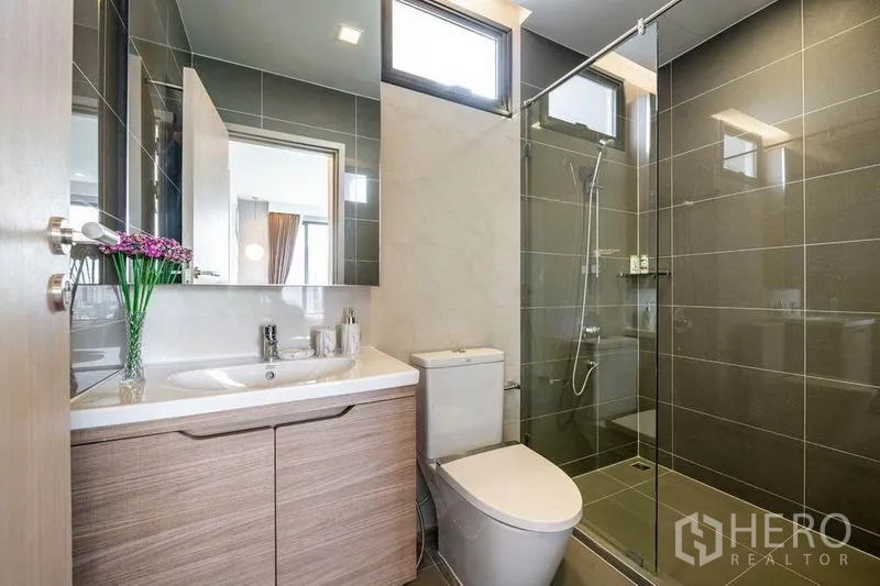 Condominium for rent or sale in Watthana, Bangkok - Second bathroom with glass shower, vanity, window and gray wall tiles.