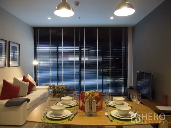 Condominium for rent or sale in Watthana, Bangkok - Open-plan living and dining room with sofa, TV and floor-to-ceiling windows with dark blinds.