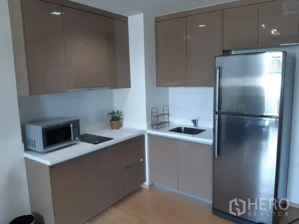 公寓 出租在Khlong Toei, Bangkok - 转角厨房，亮面棕色柜体、白色台面，配不锈钢冰箱与微波炉。