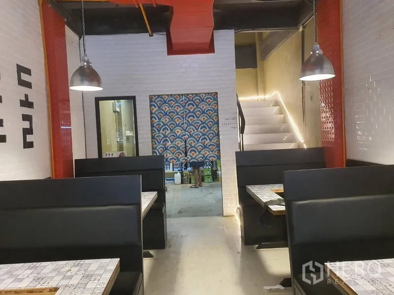 Shophouse for rent in Khlong Toei, Bangkok - Ground-floor dining area with black booths facing a rear prep zone and stairway, white tile walls and pendant lamps.