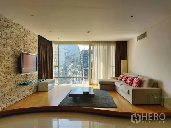 Condominium for rent in Watthana, Bangkok - Living room with stone accent wall, sofa, mounted TV, and wide city‑view windows.
