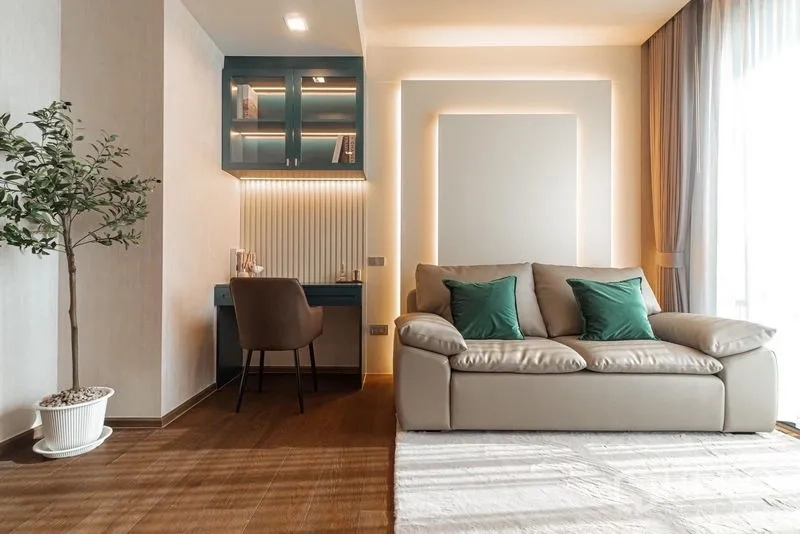 Condominium for rent in Khlong Toei, Bangkok - Living area with a beige sofa, green cushions, and a study nook by the window.