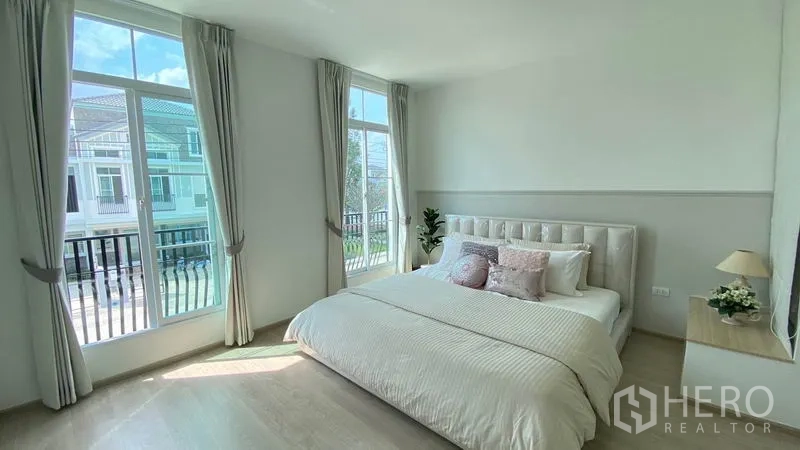 Townhouse for rent in Prawet, Bangkok - Sunlit master bedroom with large windows, balcony railings, curtains and soft neutral tones.
