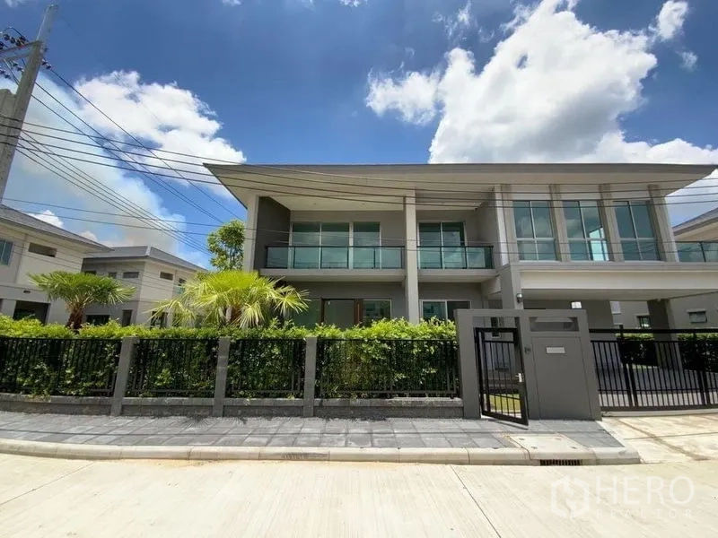 Townhouse for rent in Prawet, Bangkok - Modern two-storey townhouse exterior with glass balconies, front gate and landscaped garden under blue sky.