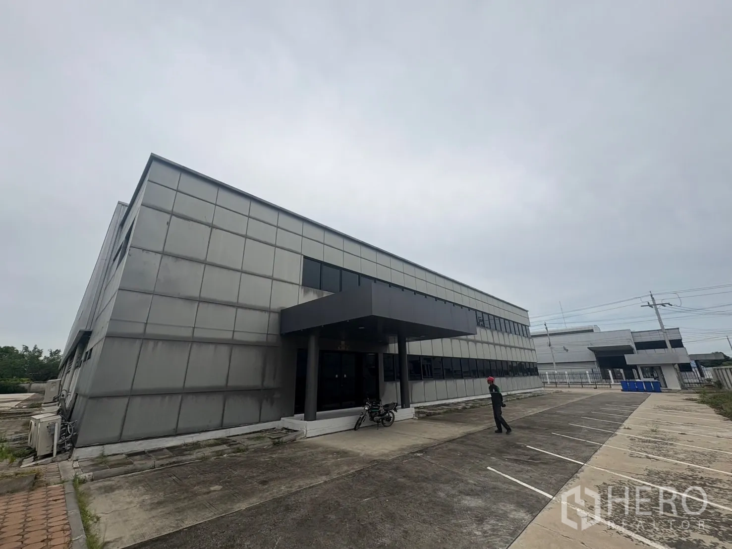 Factory for rent or sale in Samut Prakan - Back view of the site with office wing, shrubs, and marked parking grid.