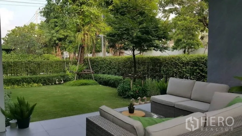 Detached House for rent or sale in Prawet, Bangkok - Covered patio with outdoor sofa set facing a lush private lawn and hedged garden.