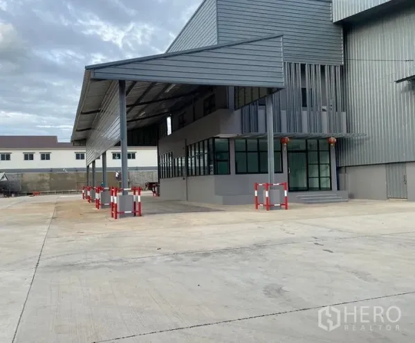Warehouse for rent in Bang Phli, Samut Prakan - Side view of warehouse canopy and office windows with ample truck maneuvering space and red‑white safety bollards.