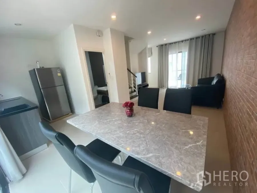 Townhouse for rent or sale in Bang Kapi, Bangkok - Open-plan dining and living area with a marble table, brick feature wall, modern pantry kitchen and staircase, filled with natural light.