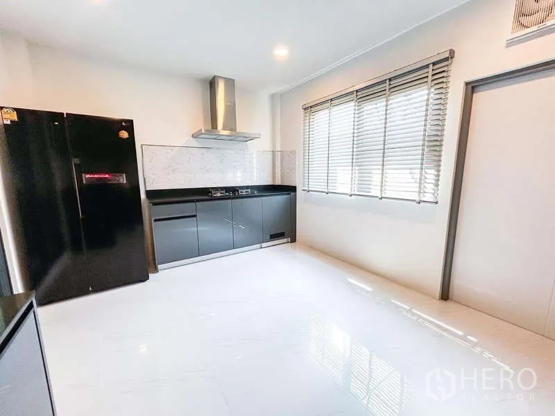 Detached House for rent in Bang Phli, Samut Prakan - Bright kitchen with black cabinetry, marble backsplash, range hood and large black fridge beside a window.