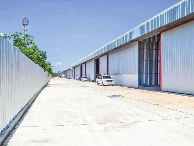 Warehouse for rent in Bang Phli, Samut Prakan - Row of metal‑clad warehouse units with a wide concrete driveway for truck access in Bang Phli under a clear sky.