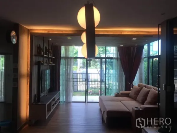 บ้าน ขายในPak Kret, Nonthaburi - ห้องนั่งเล่นอบอุ่น ผนังกระจกเต็มบาน โคมไฟแขวน และโซฟาเข้ามุมเชื่อมต่อสวน