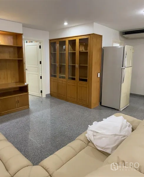 Condominium for rent or sale in Watthana, Bangkok - Bright living room with beige leather sectional, glass display cabinet, built-in shelving, split A/C and a large refrigerator on polished stone floors.
