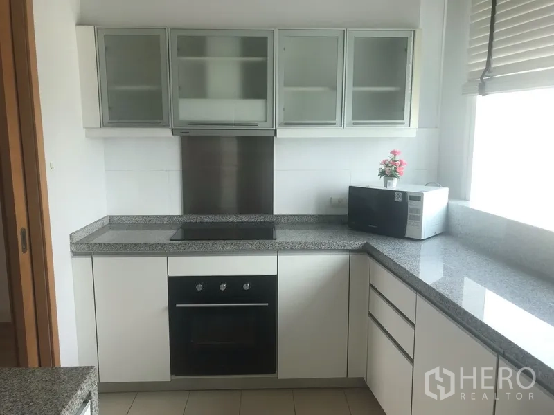 Condominium for rent in Khlong Toei, Bangkok - Modern closed kitchen with white cabinets, granite countertops, oven, cooktop, and microwave by a window.