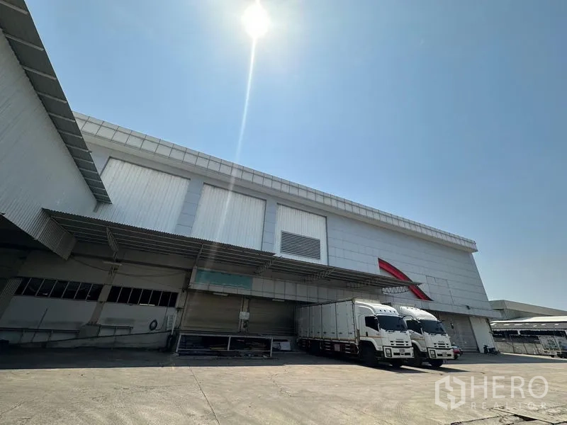 Warehouse for rent in Lam Luk Ka, Pathum Thani - Wide warehouse facade with multiple loading bays and two trucks at the docks.