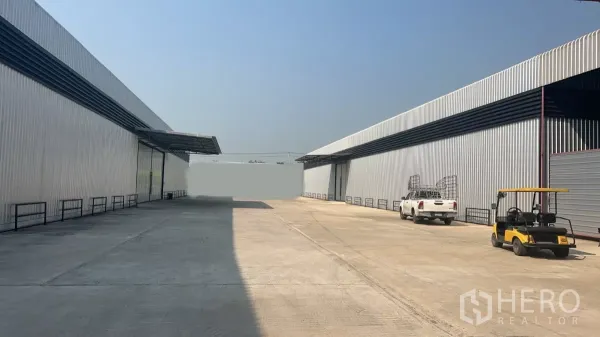 货栈 出租在Lat Lum Kaeo, Pathum Thani - 封闭园区内两侧金属板仓库与宽阔混凝土通道，可见车辆与带雨棚装卸区