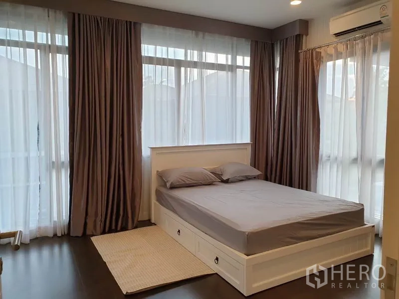 Detached House for rent in Bang Kapi, Bangkok - Corner bedroom with large wraparound windows, sheer curtains, air conditioner, and storage-bed frame.