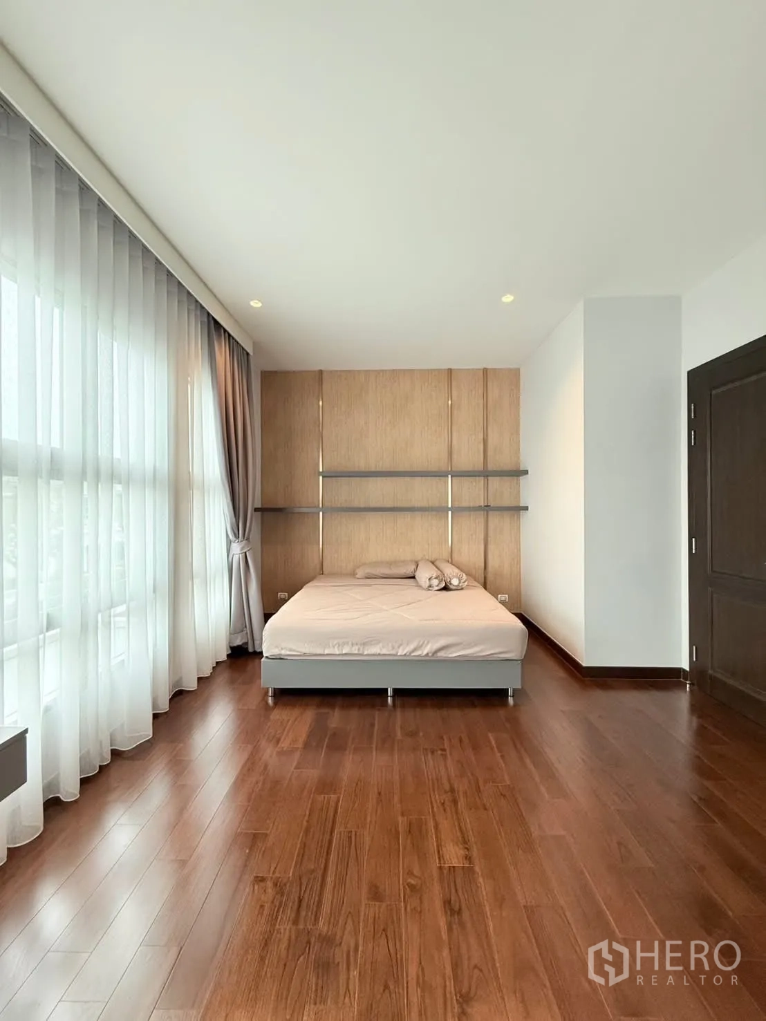 独立式住宅 出租或出售在Bang Phli, Samut Prakan - 宽敞卧室，全景大窗、木地板与简约床和木质背景墙。