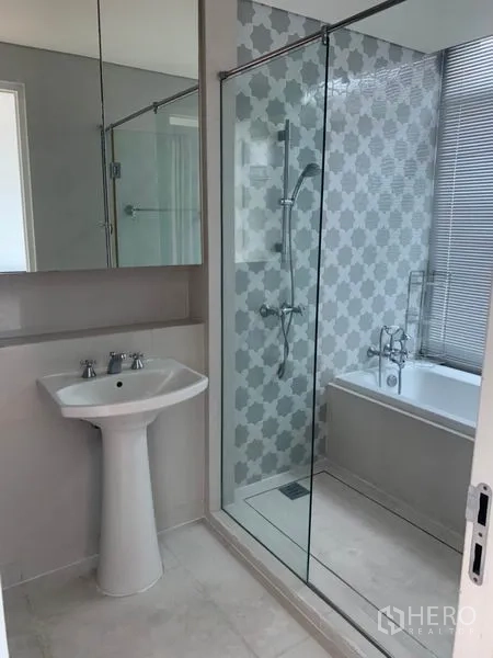 Condominium for rent in Khlong Toei, Bangkok - Contemporary bathroom with glass-enclosed shower, separate bathtub with patterned tile and a pedestal sink.
