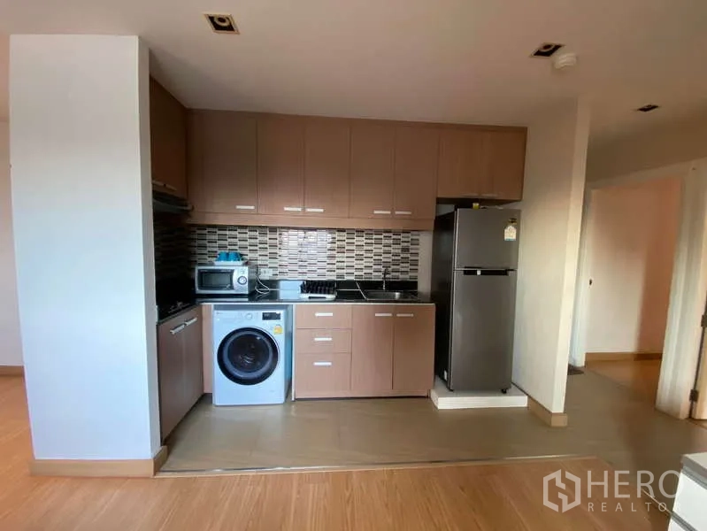 Condominium for rent in Khlong Toei, Bangkok - Compact fitted kitchen with brown cabinets, mosaic tile backsplash, cooktop, microwave, fridge and washing machine.