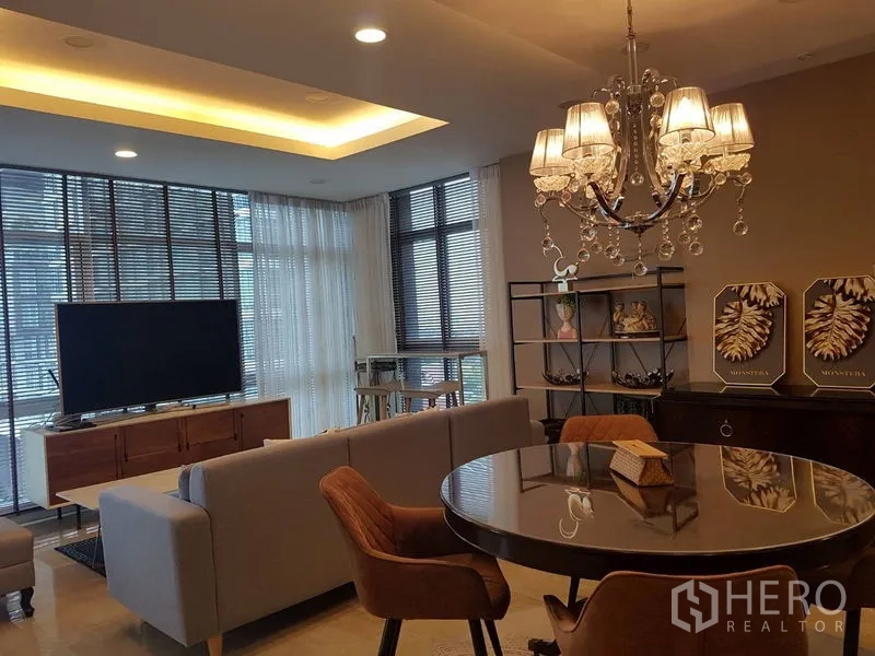 公寓 出租或出售在Bang Na, Bangkok - 餐区圆桌与水晶吊灯，旁连大窗户的起居区与电视。