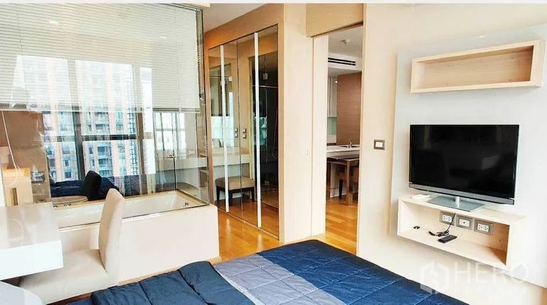 Condominium for rent in Ratchathewi, Bangkok - Bedroom angle with wall‑mounted TV, glass partition to bathroom, built‑in desk and wood floors.