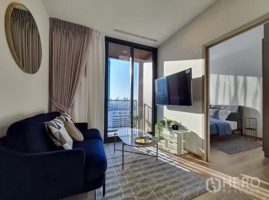 Condominium for rent in Khlong Toei, Bangkok - Sunlit living room with navy sofa, wall‑mounted TV and balcony city view.