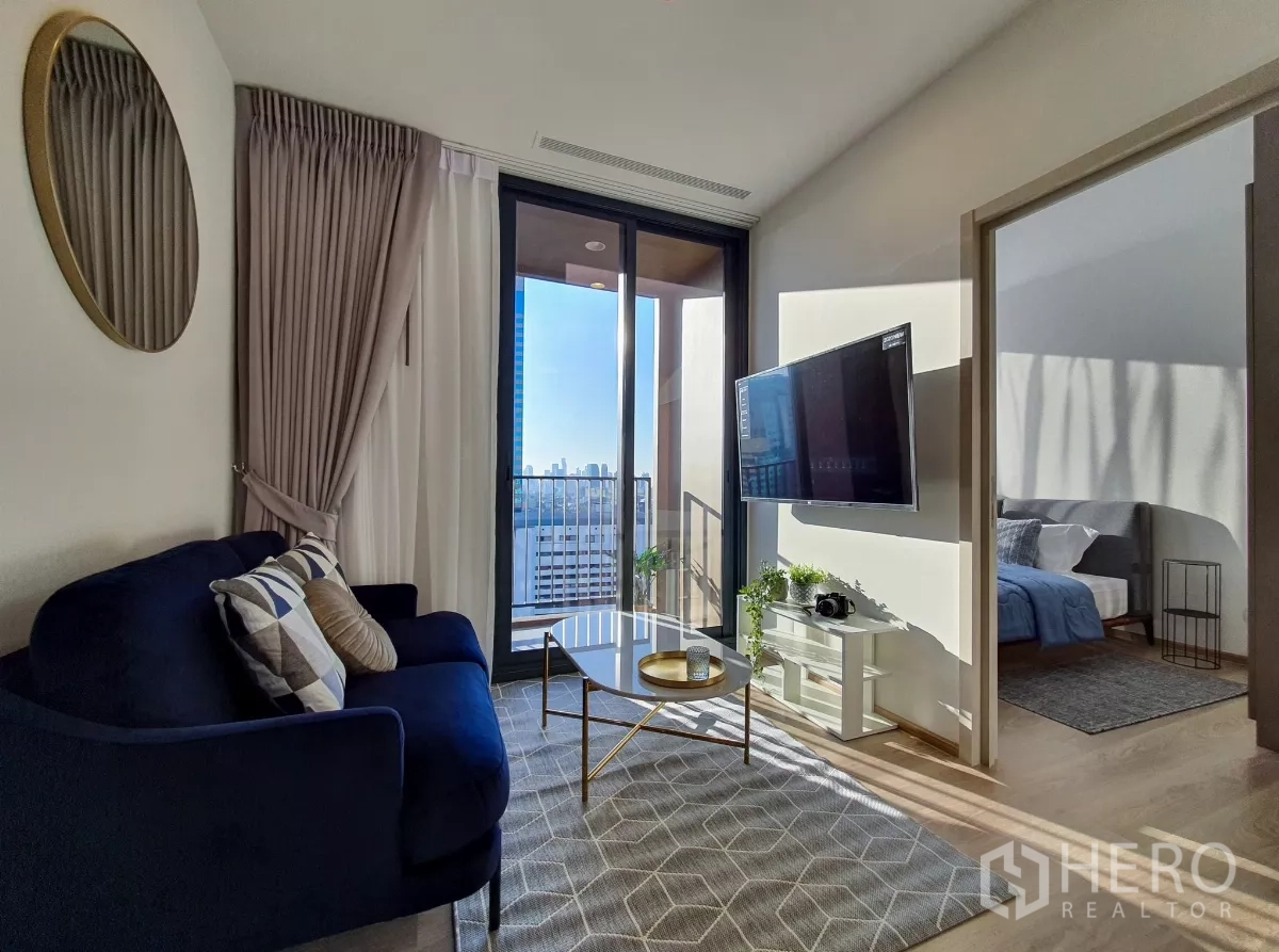 Condominium for rent in Khlong Toei, Bangkok - Sunlit living room with navy sofa, wall‑mounted TV and balcony city view.