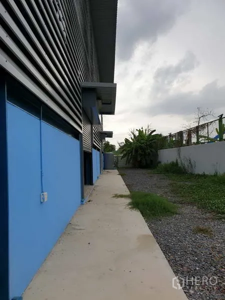 Factory for sale in Sai Noi, Nonthaburi - Concrete walkway and gravel strip running beside the factory with small awnings and greenery beyond the wall.