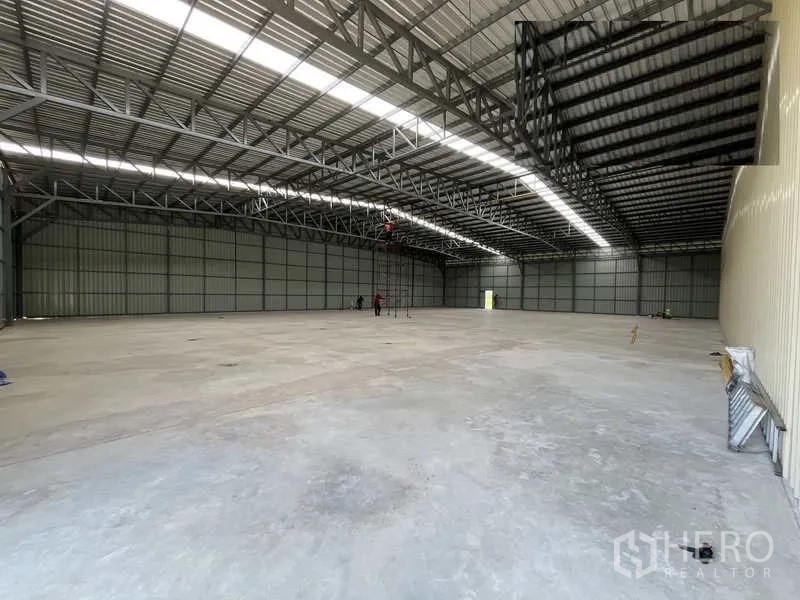 Warehouse for rent or sale in Pak Kret, Nonthaburi - Large warehouse interior with workers setting up utilities beneath continuous roof skylights.
