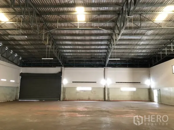 Factory for rent in Nonthaburi - Spacious warehouse interior with a high steel truss ceiling, lighting and a roller door.