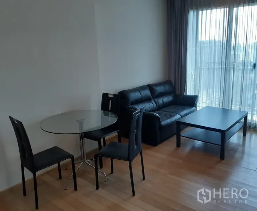 Condominium for rent in Khlong Toei, Bangkok - Bright living and dining area with black leather sofa, glass table, wood floors and floor-to-ceiling window opening to a balcony.