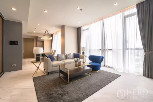Condominium for rent in Watthana, Bangkok - Sunlit living room with floor-to-ceiling sheer curtains, cream sofa, blue accent chair, gold coffee table and chandelier.