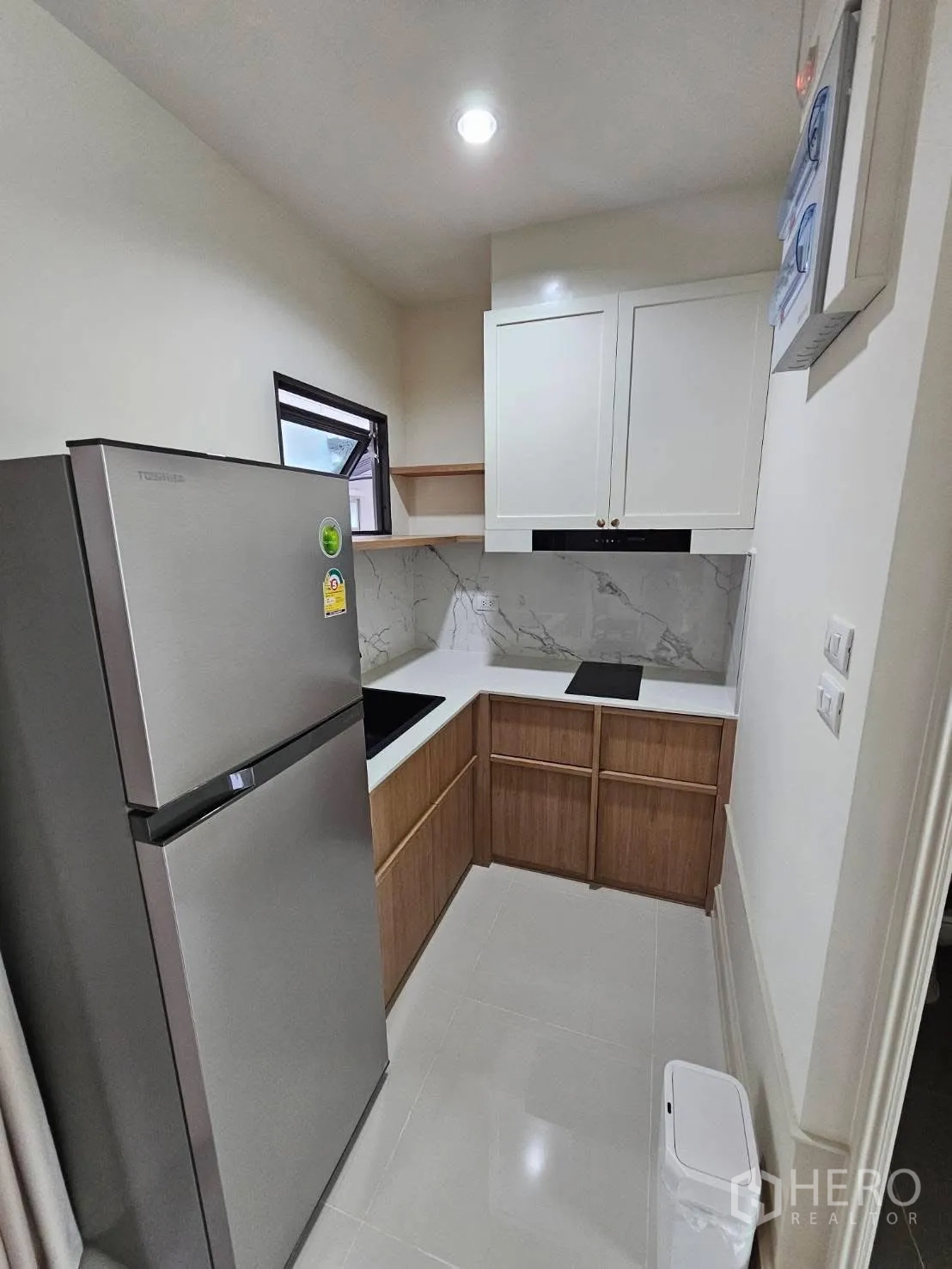 Townhouse for rent in Saphan Sung, Bangkok - Compact L-shaped kitchen with white and wood cabinets, marble-look backsplash, induction hob, and stainless refrigerator.