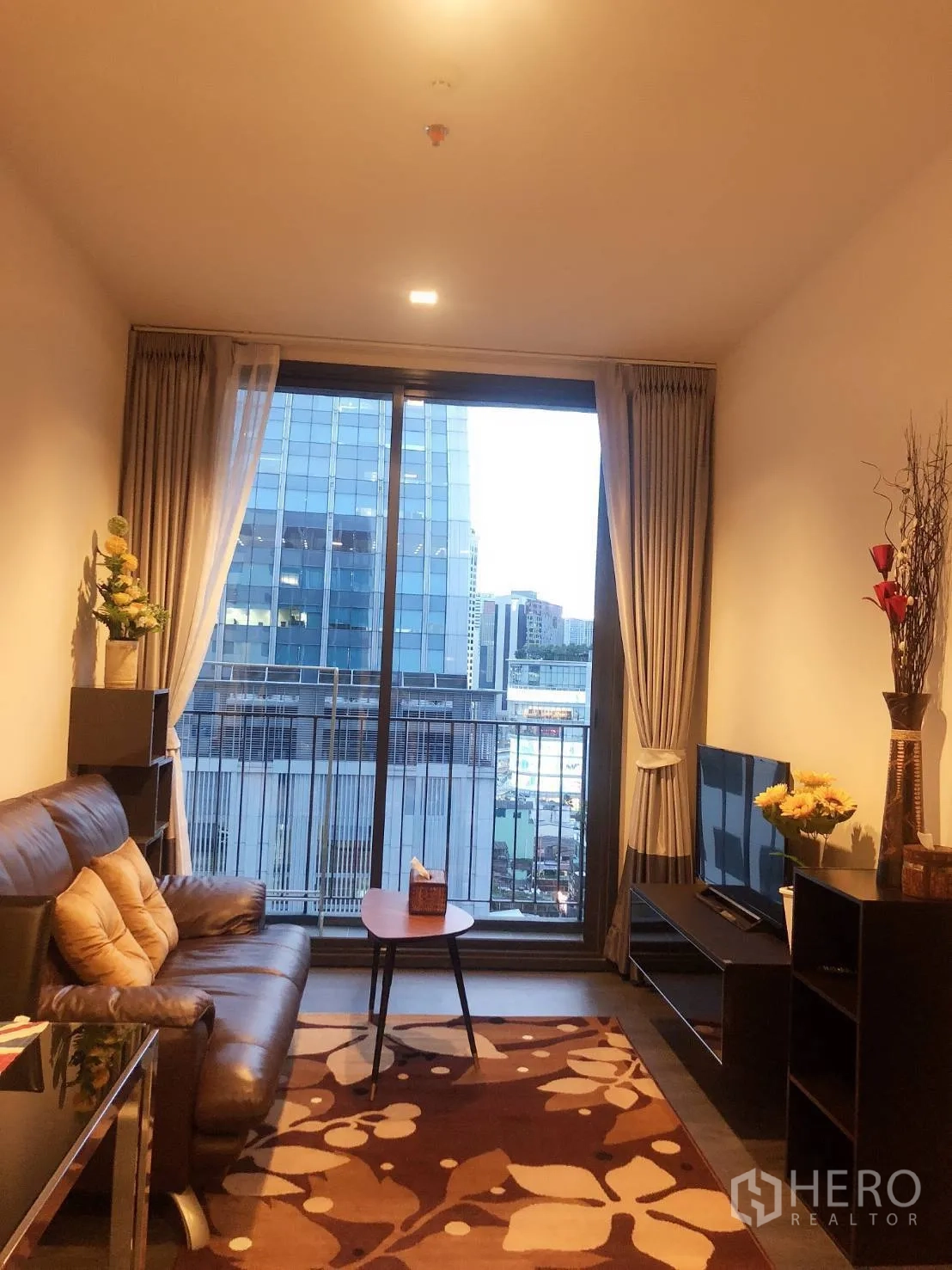คอนโด ให้เช่าในWatthana, Bangkok - ห้องนั่งเล่นโซฟาหนัง ชั้นวางทีวี พรม และประตูกระจกเต็มบานออกสู่ระเบียงวิวเมือง