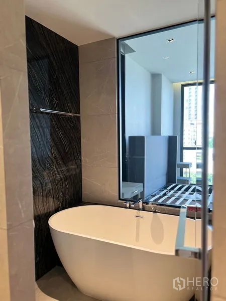 Condominium for sale in Watthana, Bangkok - Contemporary bathroom featuring a freestanding oval bathtub, marble‑style accent wall and large window looking toward the bedroom and city view.