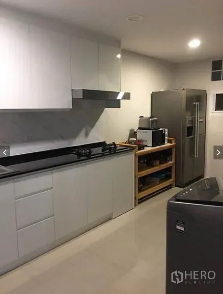 Townhouse for rent or sale in Suan Luang, Bangkok - Modern kitchen with white cabinetry, black countertop, gas hob, hood, and stainless refrigerator.