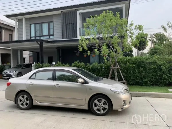 独立式住宅 出租或出售在Samut Prakan - 现代两层独栋别墅外观，带车棚、阳台与绿篱前庭。