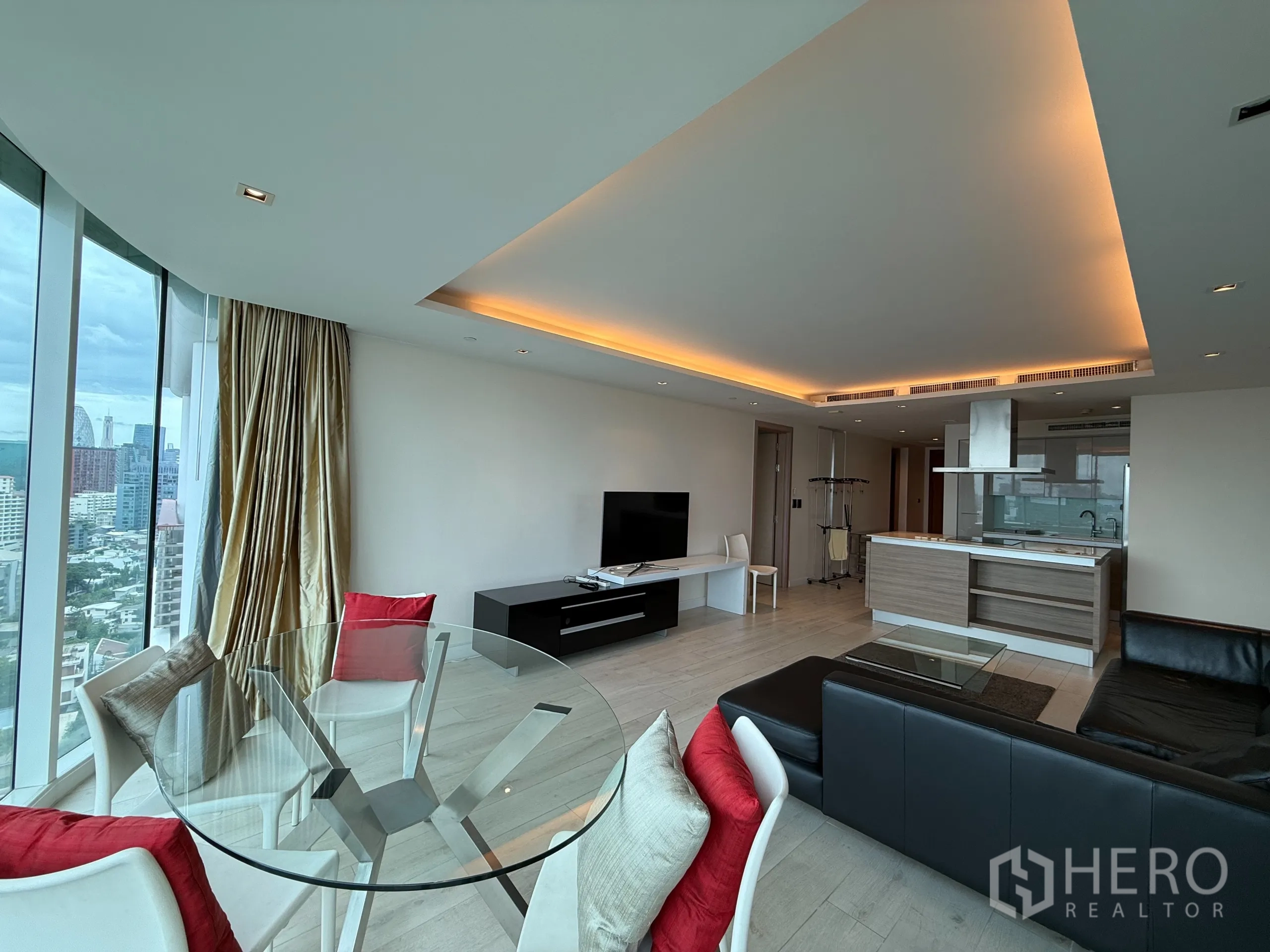 Condominium for rent in Phaya Thai, Bangkok - Open‑plan living and dining with glass table, black sofa, TV unit, island kitchen and floor‑to‑ceiling windows showing Bangkok skyline.