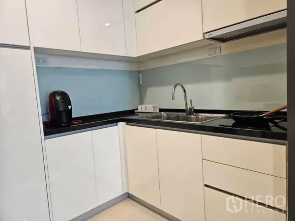 Condominium for rent or sale in Khlong Toei, Bangkok - Modern L-shaped kitchen with glossy white cabinets, black countertop, glass backsplash, sink and cooktop.