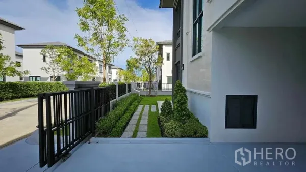 บ้าน ขายในSamut Prakan - สวนหน้าบ้านพร้อมประตูรั้วเหล็ก พุ่มไม้ตัดแต่ง และทางเดินข้างตัวบ้านสมัยใหม่