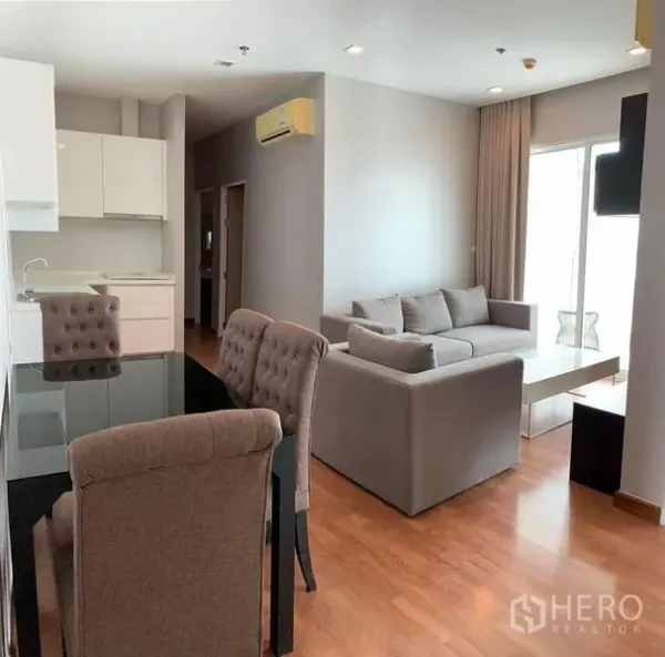 Condominium for rent or sale in Bang Na, Bangkok - Open-plan living and dining space with modern kitchenette, gray sofa set, dining table and balcony access.