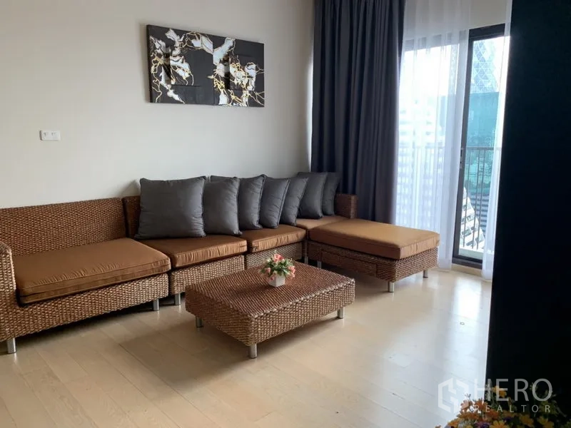 Condominium for rent in Phaya Thai, Bangkok - Bright living room with rattan sectional sofa, coffee table, light wood floors, and a balcony door with curtains.