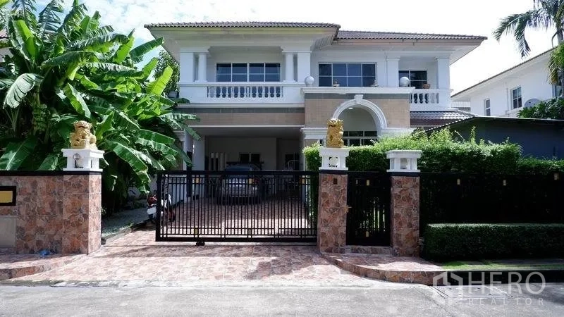 独立式住宅 出租在Prawet, Bangkok - 两层独栋别墅外立面，带围栏电动门、铺砖车道与有顶车棚，绿意环绕。
